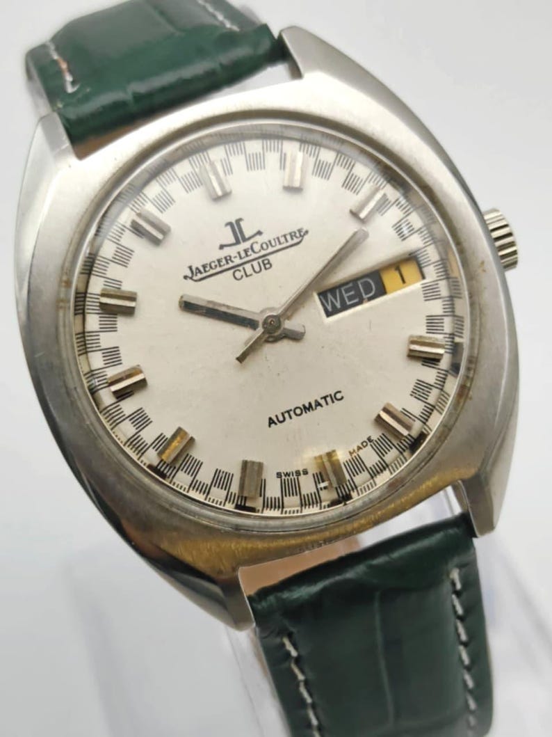 Puede incluir: Un reloj autom&aacute;tico Jaeger-LeCoultre Club vintage con esfera plateada, visualizaci&oacute;n de d&iacute;a y fecha y correa de cuero verde. El reloj tiene una caja de acero inoxidable y presenta el texto "Jaeger-LeCoultre Club" y "Automatic".