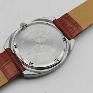 May include: A vintage wristwatch with a silver-colored case and a brown leather strap. The watch face is not visible. The back of the watch has a circular pattern. The watch strap is 0.75 inches wide.