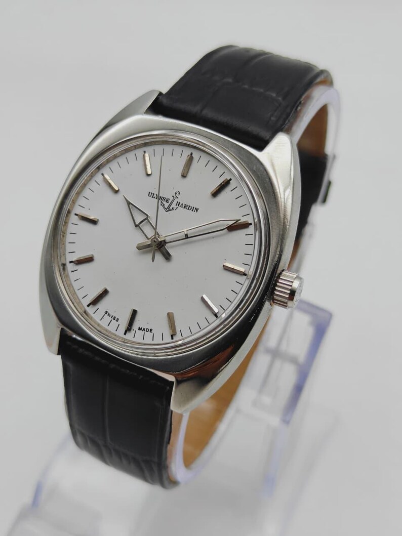 May include: A silver-toned wristwatch with a white face and black leather band. The watch face features the brand name "ULYSSE NARDIN" and the words "SWISS MADE". The watch has a classic design.