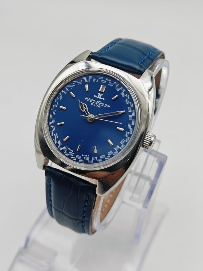 Puede incluir: Un reloj Jaeger-LeCoultre Club vintage con esfera azul y caja plateada. El reloj presenta una correa de cuero azul y el texto "JAEGER-LECOULTRE CLUB" en la esfera. El reloj se exhibe sobre un soporte transparente.