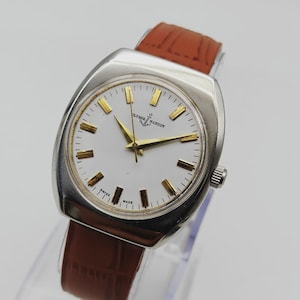 May include: A vintage Ulysse Nardin wristwatch with a silver-toned case and a white dial. The watch features gold-toned hour markers and hands, and a brown leather strap. The watch is marked "Swiss Made".