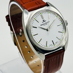 May include: A silver-toned Jaeger-LeCoultre Club watch with a white face and brown leather band. The watch face features black and green markers and the Jaeger-LeCoultre logo. The band is a rich, dark brown color.