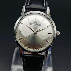 Op de afbeelding: Een zilverkleurig Jaeger-LeCoultre Club horloge met een zwarte leren band. De wijzerplaat heeft een geborstelde afwerking en is voorzien van de merknaam en de tekst "Swiss Made". Het horloge wordt op een transparante standaard getoond.