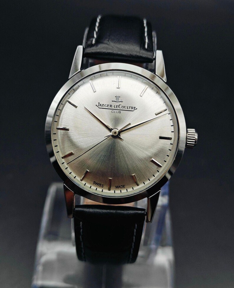 Op de afbeelding: Een zilverkleurig Jaeger-LeCoultre Club horloge met een zwarte leren band. De wijzerplaat heeft een geborstelde afwerking en is voorzien van de merknaam en de tekst "Swiss Made". Het horloge wordt op een transparante standaard getoond.