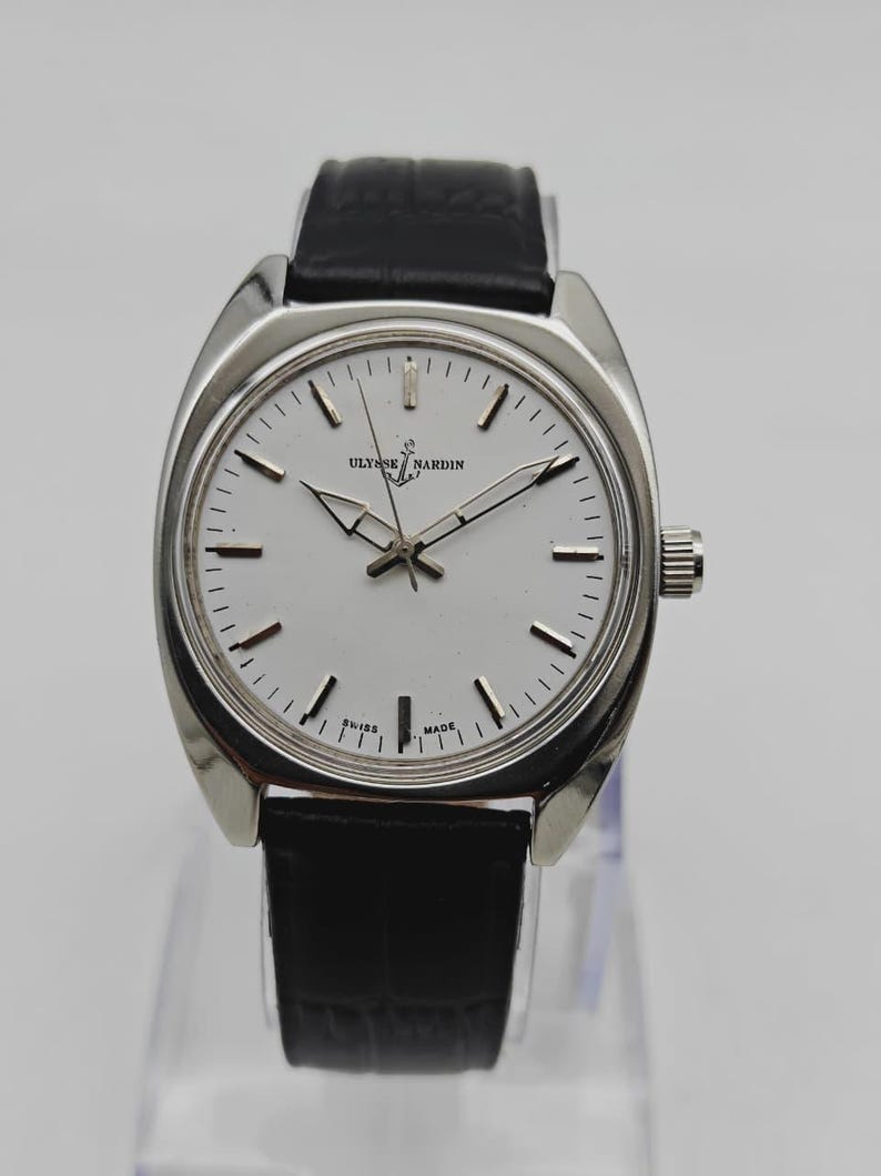 May include: A silver-toned Ulysse Nardin wristwatch with a white face and black leather band. The watch has a round face with silver hands and markers. The watch is displayed on a clear stand.