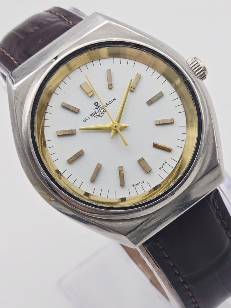 Pu&ograve; includere: Un orologio da polso vintage argentato con quadrante bianco e dettagli dorati. L'orologio presenta un cinturino in pelle marrone e il marchio "Ulysse Nardin" sul quadrante. Le lancette e gli indici sono dorati.
