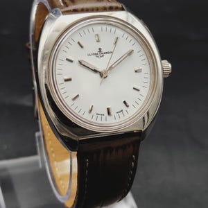May include: A Ulysse Nardin wristwatch featuring a round silver-tone case, a white dial with silver-tone hour markers, and silver-tone hands. The watch includes a brown leather strap with a textured finish and white stitching along the edges.