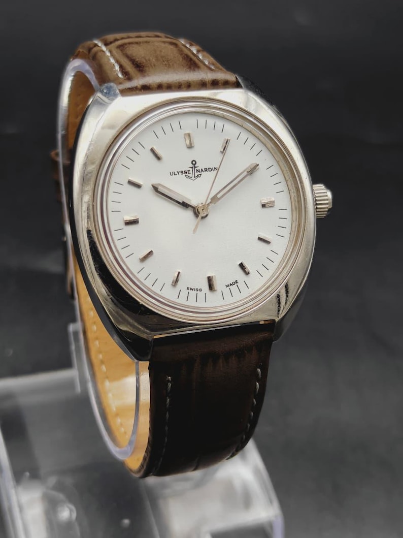 May include: A Ulysse Nardin wristwatch featuring a round silver-tone case, a white dial with silver-tone hour markers, and silver-tone hands. The watch includes a brown leather strap with a textured finish and white stitching along the edges.