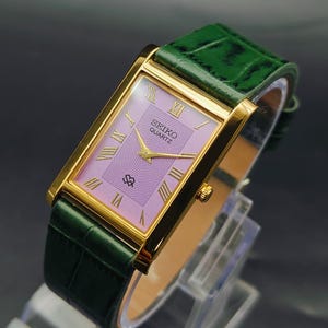 May include: A rectangular Seiko Quartz wristwatch featuring a gold-tone case, a light purple dial with a textured center, and gold Roman numeral hour markers. The watch is paired with a dark green leather strap with a crocodile-embossed pattern.