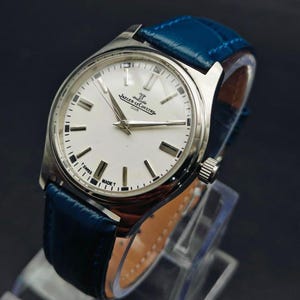 Peut inclure: Une montre Jaeger-LeCoultre Club argent&eacute;e avec un cadran blanc et un bracelet en cuir bleu. Le cadran comporte des marqueurs et des aiguilles noirs, avec le nom de la marque et le texte "SWISS MADE T". Le bracelet mesure environ 20 mm de large.