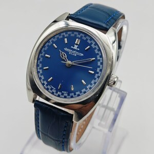 Puede incluir: Un reloj Jaeger-LeCoultre Club vintage con esfera azul y caja plateada. El reloj presenta una correa de cuero azul y el texto "JAEGER-LECOULTRE CLUB" en la esfera. El reloj se exhibe sobre un soporte transparente.