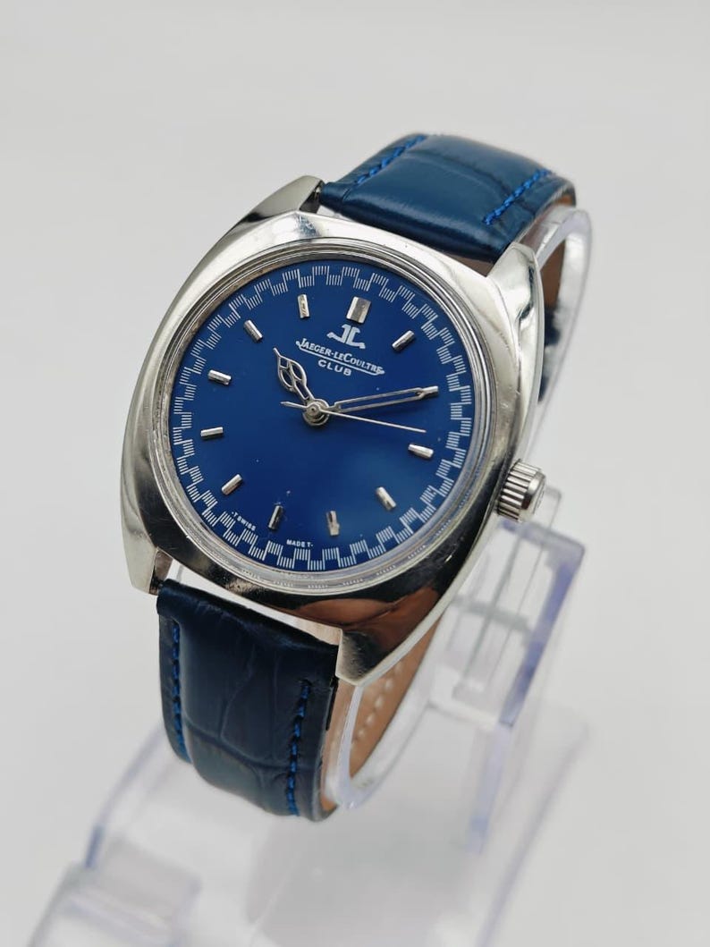 Puede incluir: Un reloj Jaeger-LeCoultre Club vintage con esfera azul y caja plateada. El reloj presenta una correa de cuero azul y el texto "JAEGER-LECOULTRE CLUB" en la esfera. El reloj se exhibe sobre un soporte transparente.