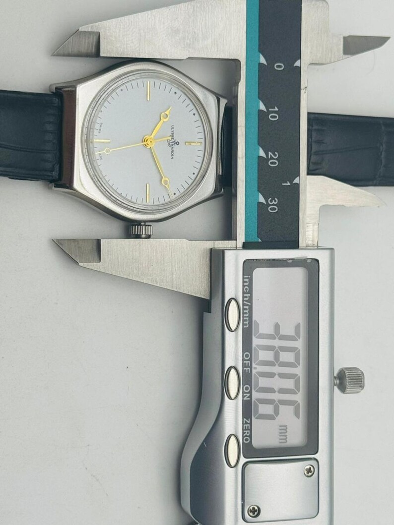 May include: A silver-toned wristwatch with a light-colored face and gold hands is measured with a digital caliper. The watch has a black leather band. The caliper displays a measurement of 38.06 mm.