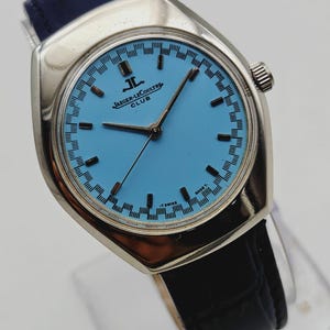 May include: A vintage Jaeger-LeCoultre Club watch with a light blue face and silver case. The watch has black hands and markers, a black leather band, and a blue leather strap. The watch face has a patterned border and the Jaeger-LeCoultre logo.