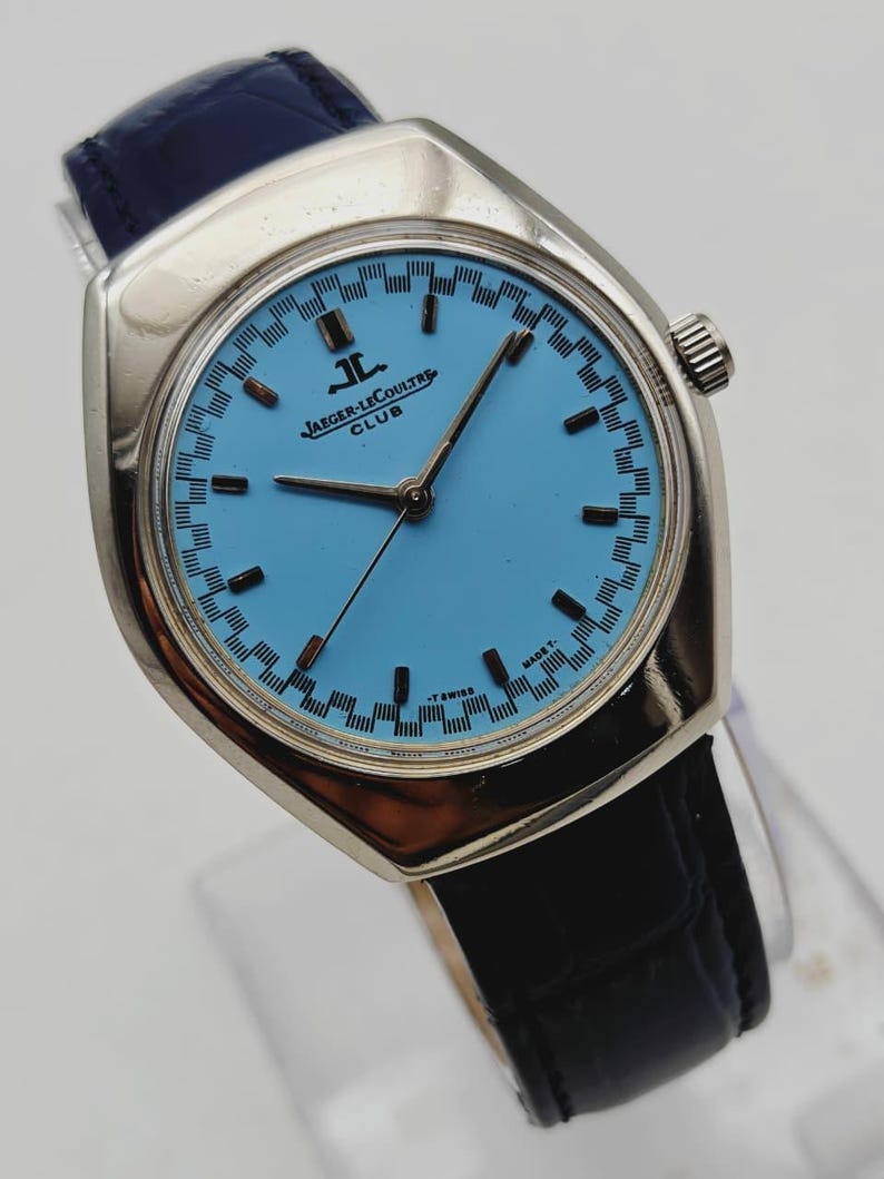 May include: A vintage Jaeger-LeCoultre Club watch with a light blue face and silver case. The watch has black hands and markers, a black leather band, and a blue leather strap. The watch face has a patterned border and the Jaeger-LeCoultre logo.