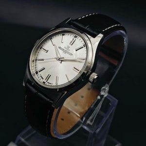 May include: A silver-toned wristwatch with a black leather band. The watch face is silver with black hour markers and hands. The band is stitched with white thread. The watch is displayed on a clear stand.
