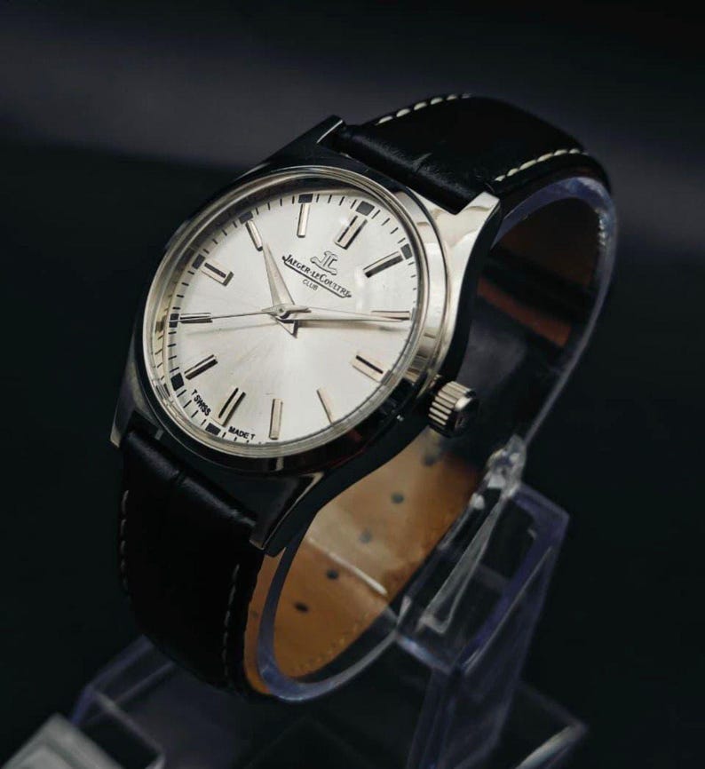 May include: A silver-toned wristwatch with a black leather band. The watch face is silver with black hour markers and hands. The band is stitched with white thread. The watch is displayed on a clear stand.