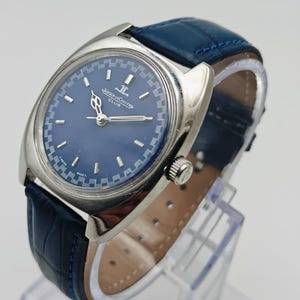 Puede incluir: Un reloj Jaeger-LeCoultre Club vintage con esfera azul y caja plateada. El reloj presenta una correa de cuero azul con un patr&oacute;n de cocodrilo. El reloj se exhibe sobre un soporte transparente.