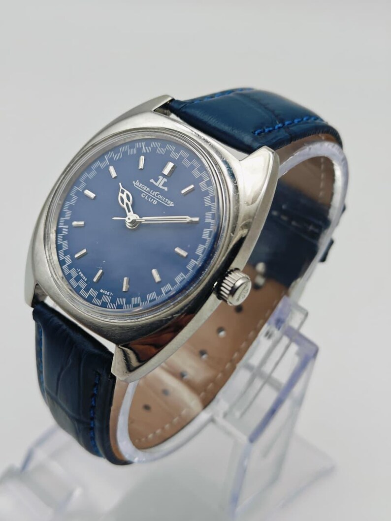 Puede incluir: Un reloj Jaeger-LeCoultre Club vintage con esfera azul y caja plateada. El reloj presenta una correa de cuero azul con un patr&oacute;n de cocodrilo. El reloj se exhibe sobre un soporte transparente.