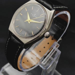 May include: A vintage Ulysse Nardin wristwatch featuring a round dark grey dial with gold-tone hands and hour markers, housed in a silver-tone metal case. The watch is attached to a black leather strap with white stitching and a light tan lining.