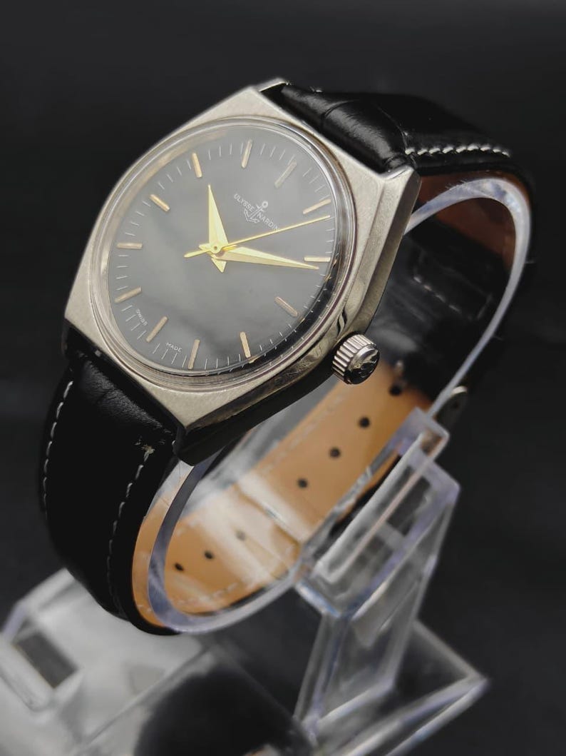 May include: A vintage Ulysse Nardin wristwatch featuring a round dark grey dial with gold-tone hands and hour markers, housed in a silver-tone metal case. The watch is attached to a black leather strap with white stitching and a light tan lining.