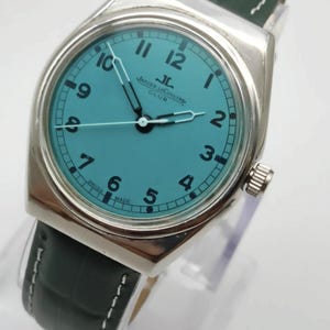 Pode incluir: Um rel&oacute;gio de pulso prateado com mostrador turquesa e algarismos pretos. O rel&oacute;gio apresenta uma bracelete em pele verde escura com pespontos brancos. O mostrador exibe a marca "Jaeger LeCoultre Club".