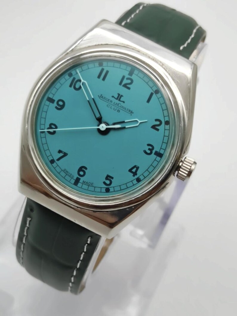 Pode incluir: Um rel&oacute;gio de pulso prateado com mostrador turquesa e algarismos pretos. O rel&oacute;gio apresenta uma bracelete em pele verde escura com pespontos brancos. O mostrador exibe a marca "Jaeger LeCoultre Club".