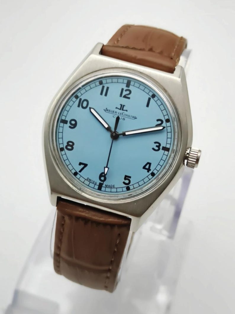 K&ouml;nnte beinhalten: Eine silberfarbene Armbanduhr mit hellblauem Zifferblatt und braunem Lederarmband. Das Zifferblatt zeigt schwarze Ziffern und Zeiger, mit dem Markennamen "Jaeger-LeCoultre". Ein klassisches Uhrendesign.
