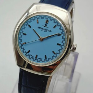 May include: A vintage Jaeger-LeCoultre Club watch with a light blue face and silver case. The watch has a dark blue leather strap. The watch face features the Jaeger-LeCoultre logo and text. The watch is displayed on a clear stand.