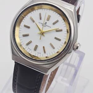 Pu&ograve; includere: Un orologio Ulysse Nardin vintage con cassa color argento, dettagli dorati e quadrante bianco. L'orologio ha un cinturino in pelle marrone. Il quadrante mostra il nome del marchio e le parole "SWISS MADE".