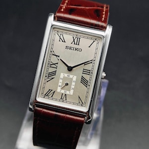 May include: A rectangular Seiko wristwatch with a silver face, black hands, and Roman numeral hour markers. The watch has a brown leather band and a silver case. The watch face includes the word "SEIKO".