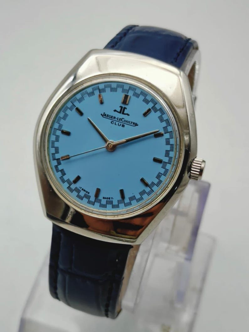 May include: A vintage Jaeger-LeCoultre Club watch with a light blue dial and silver case. The watch has a blue leather strap. The dial displays the brand name. The watch has a classic design.
