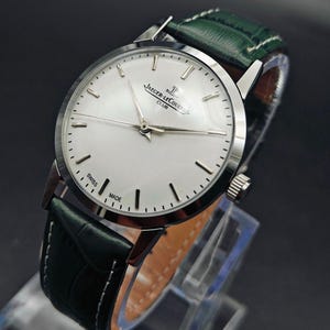 Pu&ograve; includere: Un orologio da polso argentato con quadrante bianco e indicatori neri. L'orologio presenta la scritta "JAEGER-LECOULTRE CLUB SWISS MADE" e ha un cinturino in pelle verde con cuciture bianche. L'orologio &egrave; esposto su un supporto trasparente.