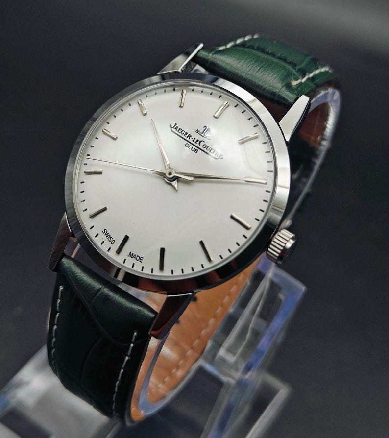 Pu&ograve; includere: Un orologio da polso argentato con quadrante bianco e indicatori neri. L'orologio presenta la scritta "JAEGER-LECOULTRE CLUB SWISS MADE" e ha un cinturino in pelle verde con cuciture bianche. L'orologio &egrave; esposto su un supporto trasparente.
