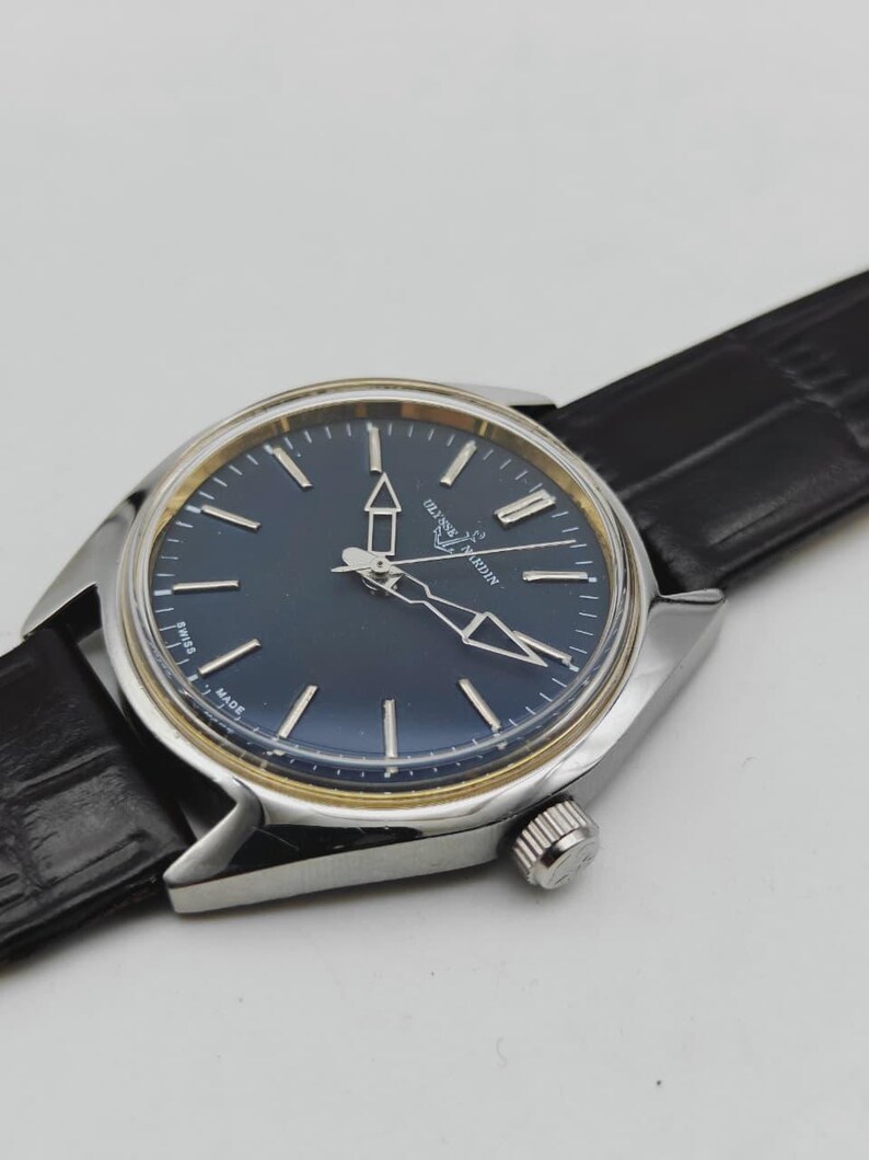 May include: A silver-toned wristwatch with a dark blue face and silver hands and markers. The watch has a black leather band. The watch face has the brand name "Ulysse Nardin" printed on it. The watch is a classic design.