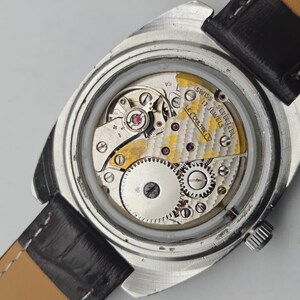 Op de afbeelding: Close-up van een vintage polshorloge met een zilverkleurige kast en een zwarte leren band. Het ingewikkelde interne mechanisme van het horloge is zichtbaar, met tandwielen, juwelen en de tekst "SWISS SEVENTEEN 17 JEWELS LE COULTRE CO."
