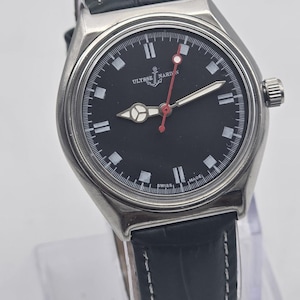 Puede incluir: Un reloj Ulysse Nardin de acero inoxidable con esfera negra, marcadores blancos y segundero rojo. El reloj tiene una correa de cuero con costuras blancas. El reloj se exhibe sobre un soporte transparente.