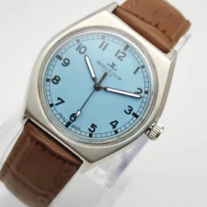 K&ouml;nnte beinhalten: Eine silberfarbene Armbanduhr mit hellblauem Zifferblatt und braunem Lederarmband. Das Zifferblatt zeigt schwarze Ziffern und Zeiger, mit dem Markennamen "Jaeger-LeCoultre". Die Uhr hat ein klassisches Design.