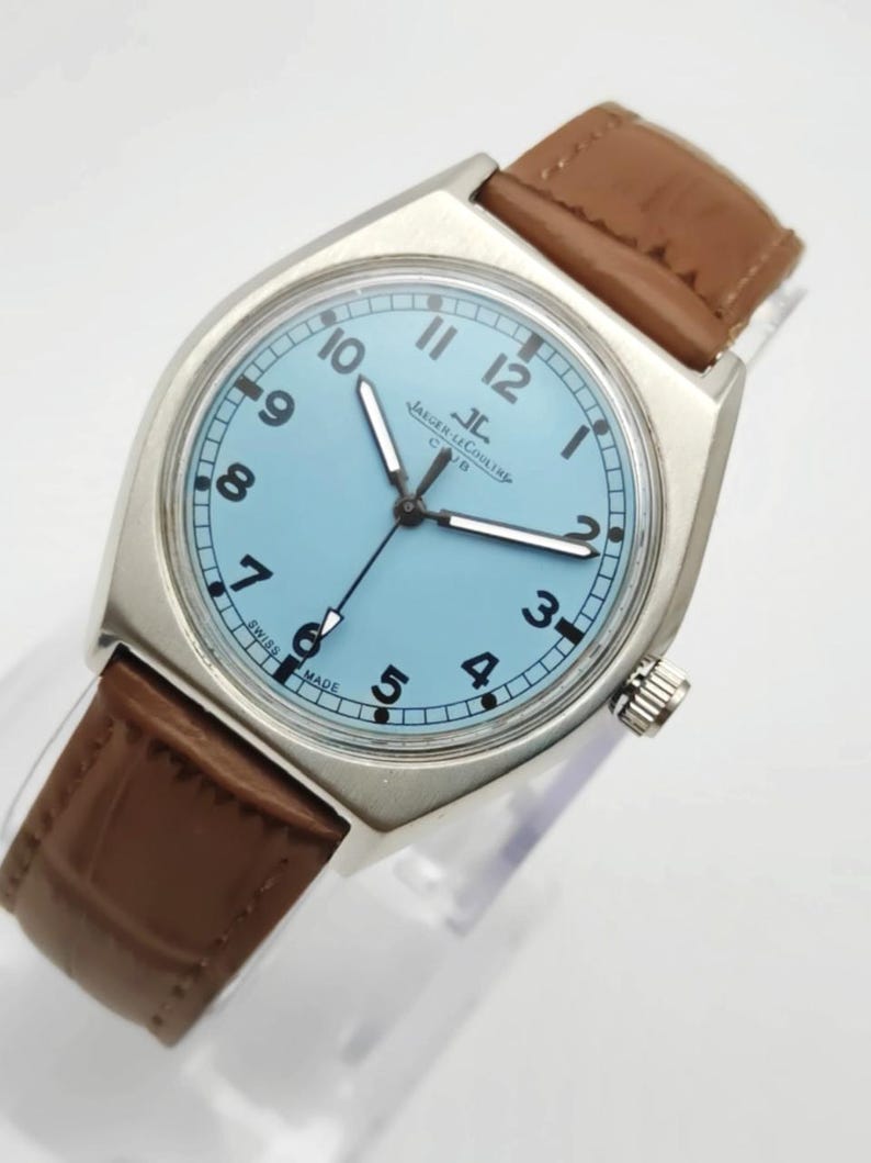 K&ouml;nnte beinhalten: Eine silberfarbene Armbanduhr mit hellblauem Zifferblatt und braunem Lederarmband. Das Zifferblatt zeigt schwarze Ziffern und Zeiger, mit dem Markennamen "Jaeger-LeCoultre". Die Uhr hat ein klassisches Design.