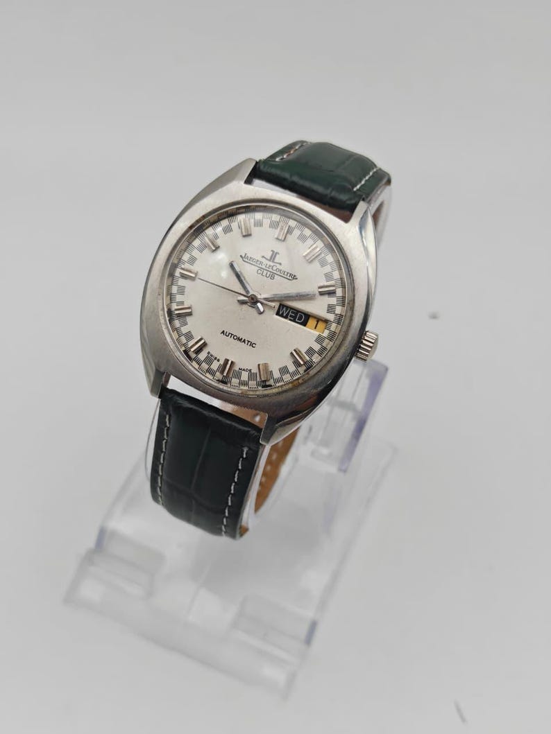 Puede incluir: Un reloj de pulsera plateado con esfera blanca, manecillas negras y visualizaci&oacute;n de d&iacute;a y fecha. El reloj tiene una correa de cuero verde. La palabra "AUTOMATIC" es visible en la esfera. El reloj se exhibe sobre un soporte transparente.