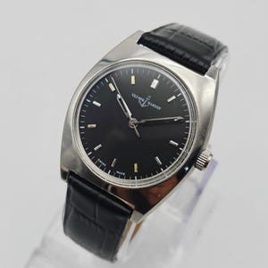 Puede incluir: Un reloj de pulsera Ulysse Nardin plateado con esfera negra y correa de cuero negro. El reloj tiene una caja rectangular y un dise&ntilde;o cl&aacute;sico. La esfera presenta marcadores de hora y agujas plateadas. La correa es de cuero negro texturizado.