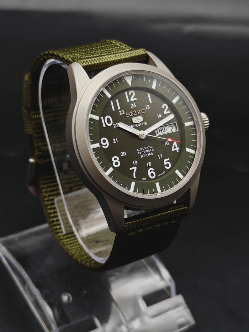 Puede incluir: Un reloj autom&aacute;tico Seiko 5 Sports con caja plateada mate y esfera verde oliva. La esfera presenta n&uacute;meros ar&aacute;bigos blancos, una ventana de d&iacute;a y fecha en la posici&oacute;n de las tres y una correa de tela de nailon verde oliva a juego.