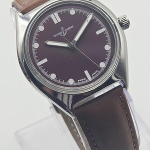 May include: A silver-toned wristwatch with a burgundy face and brown leather band. The watch face features white hour markers and hands, with the brand name "Ulysse Nardin" visible. The watch is displayed on a clear stand.