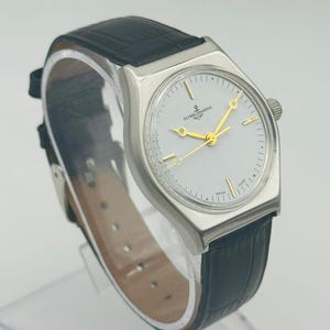May include: A silver-toned Ulysse Nardin watch with a white face and gold hands. The watch has a black leather band and a small crown. The watch face features the Ulysse Nardin logo and is set against a light background.