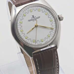 May include: A silver-toned Jaeger-LeCoultre Club watch with a white face, gold hands, and a brown leather band. The watch face features the Jaeger-LeCoultre logo and the word "CLUB". The band is approximately 0.75 inches wide.