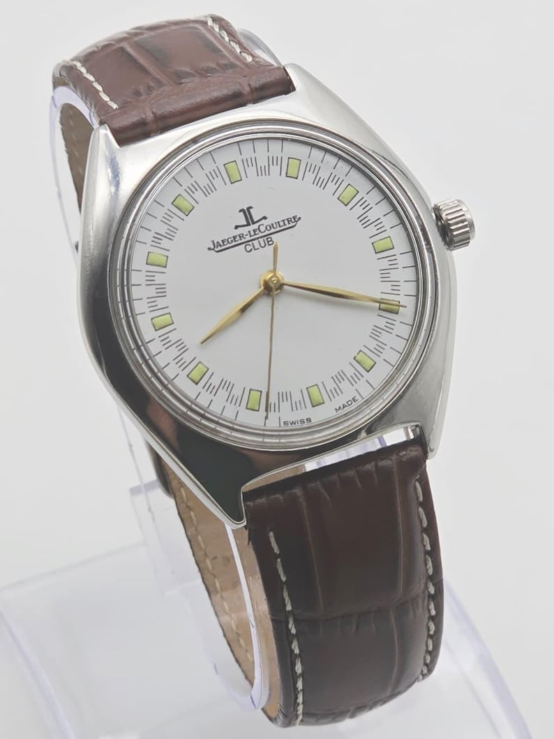 May include: A silver-toned Jaeger-LeCoultre Club watch with a white face, gold hands, and a brown leather band. The watch face features the Jaeger-LeCoultre logo and the word "CLUB". The band is approximately 0.75 inches wide.