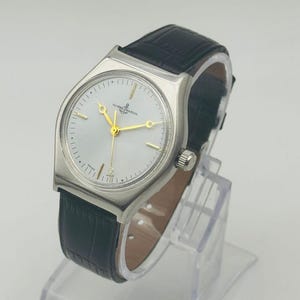May include: A silver-toned wristwatch with a white face and gold hands. The watch features the brand name "ULYSSE NARDIN" and has a black leather band. The watch is displayed on a clear stand.
