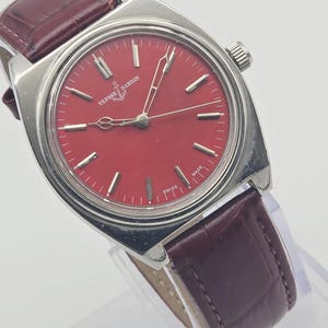 May include: A vintage Ulysse Nardin wristwatch with a vibrant red dial and silver-tone case. The watch features a burgundy leather strap. The dial has silver-tone hour markers and hands. The watch is marked "SWISS MADE".