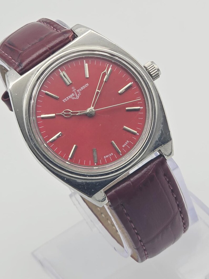 May include: A vintage Ulysse Nardin wristwatch with a vibrant red dial and silver-tone case. The watch features a burgundy leather strap. The dial has silver-tone hour markers and hands. The watch is marked "SWISS MADE".