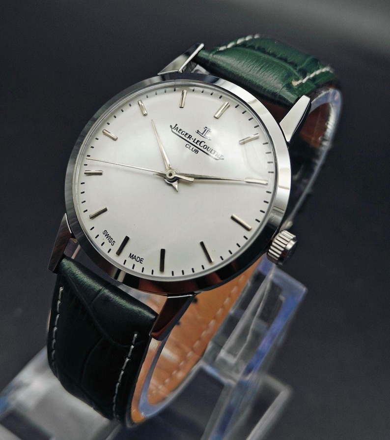 Pu&ograve; includere: Un orologio da polso argentato con quadrante bianco e indicatori neri. L'orologio presenta la scritta "JAEGER-LECOULTRE CLUB SWISS MADE" e ha un cinturino in pelle verde con cuciture bianche. L'orologio &egrave; esposto su un supporto trasparente.